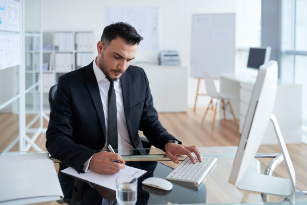 caucasian-man-business-suit-sitting-office-working-computer-writing-paper_1098-20577